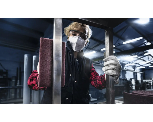 Een persoon met een veiligheidsbril en een ademhalingsmasker houdt een schuurspons vast in een industriële omgeving.