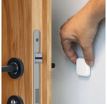 Een hand bevestigt een magnetische deurstopper aan een muur naast een houten deur met een zilverkleurig deurslot.