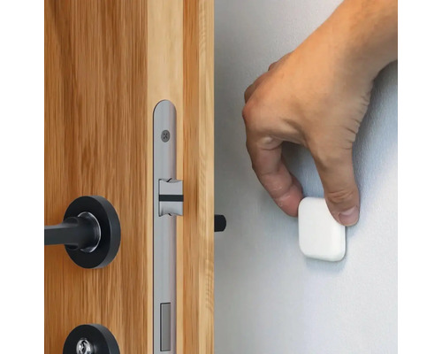 Een hand bevestigt een magnetische deurstopper aan een muur naast een houten deur met een zilverkleurig deurslot.