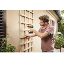 Een man bevestigt een houten trelliswand met een accuschroevendraaier aan een buitenmuur.