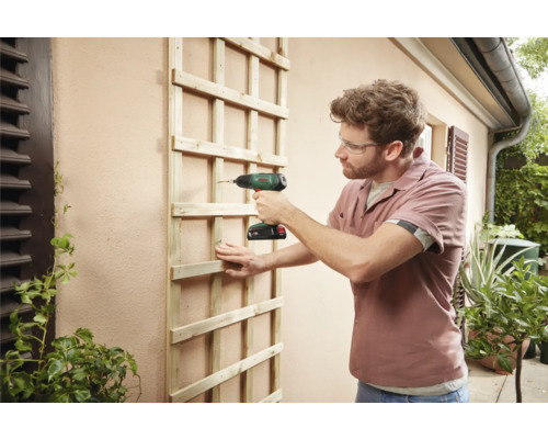 Een man bevestigt een houten trelliswand met een accuschroevendraaier aan een buitenmuur.