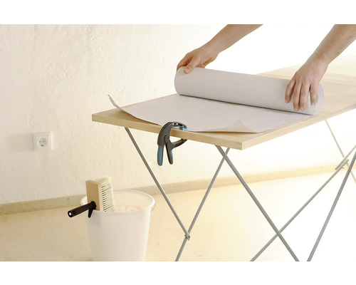 Persoon rolt behang uit op een behangtafel. Emmer en kwast staan klaar.