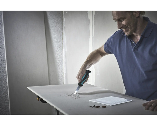 Een man zaagt met een handzaag een gat in een gipsplaat. Gereedschap en een tegel liggen op tafel.