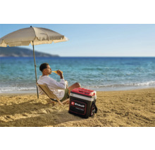 Een man ontspant op het strand onder een parasol met een Einhell koelbox naast zich