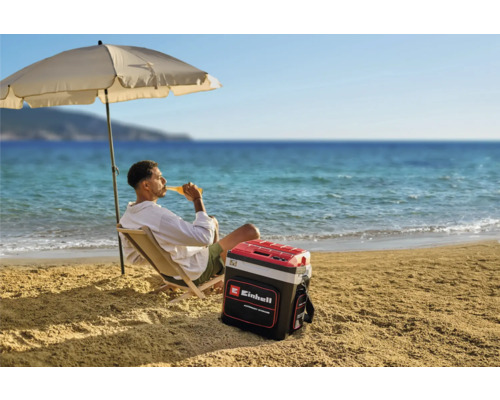 Een man ontspant op het strand onder een parasol met een Einhell koelbox naast zich