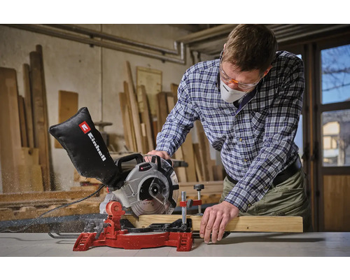 Een man met veiligheidsbril en ademhalingsmasker zaagt een houten plank met een Einhell verstekzaag.