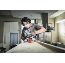 Iemand gebruikt een cirkelzaag om een houten plank te zagen, beschermd met een masker, bril en gehoorbescherming.