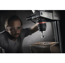 Een kolomboormachine met een man met een veiligheidsbril, die een gat boort in een stuk metaal.