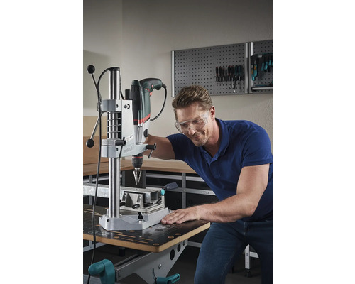 Man met veiligheidsbril boort een werkstuk met een tafelboormachine op een werkbank.