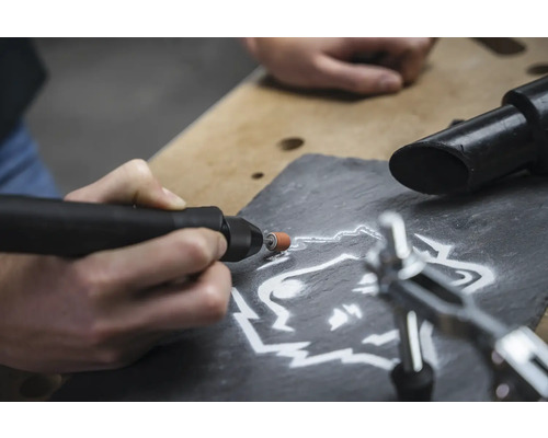 Close-up van een persoon die met een multifunctioneel gereedschap een wolfsmotief in een leisteen graveert.