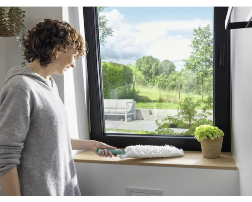 Vrouw maakt vensterbank schoon met een plumeau. Raam met uitzicht op groen.