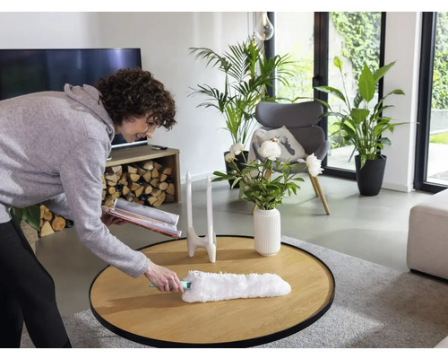 Vrouw stoffert een tafel in een lichte woonkamer met een plumeau.