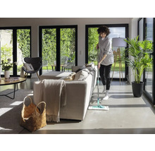 Vrouw dweilt vloer in een lichte woonkamer met grote ramen en een minimalistische inrichting.