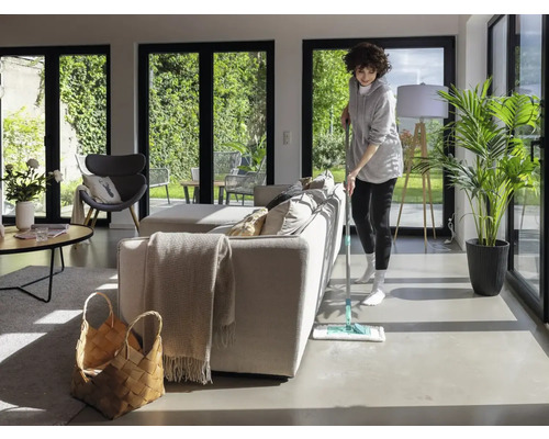 Vrouw dweilt vloer in een lichte woonkamer met grote ramen en een minimalistische inrichting.