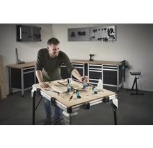 Een man werkt aan een houten frame op een werkbank in een werkplaats. Gereedschap en werkplaatsapparatuur zijn zichtbaar op de achtergrond.