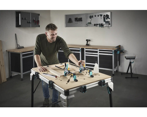Een man werkt aan een houten frame op een werkbank in een werkplaats. Gereedschap en werkplaatsapparatuur zijn zichtbaar op de achtergrond.