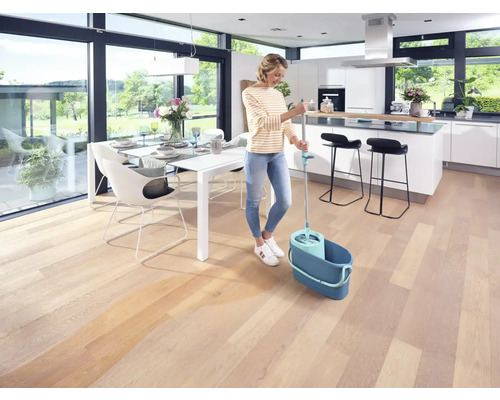 Vrouw dweilt houten vloer met dweil en emmer in een lichte woonkamer.