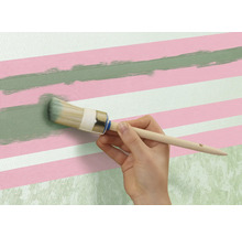 Een persoon schildert met een ronde kwast een groene lijn op een muur met roze en witte strepen