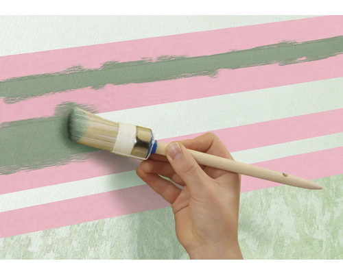 Een persoon schildert met een ronde kwast een groene lijn op een muur met roze en witte strepen