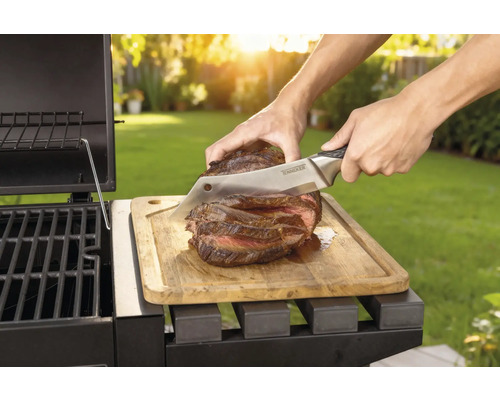 Bereide steak wordt met een mes op een houten plank naast een barbecue gesneden.