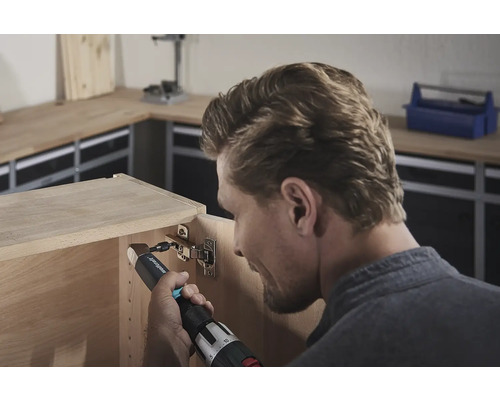 Een man bevestigt een scharnier met een accuschroevendraaier aan een houten kast.