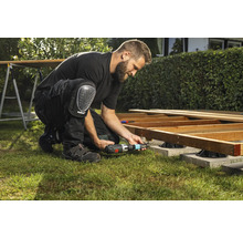 Een man bouwt een houten terras met een accuschroevendraaier en kniebeschermers in de tuin.