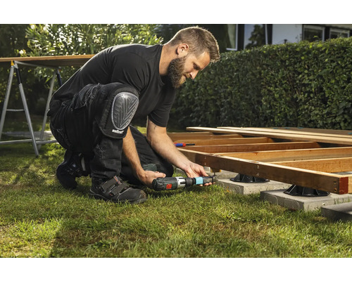 Een man bouwt een houten terras met een accuschroevendraaier en kniebeschermers in de tuin.