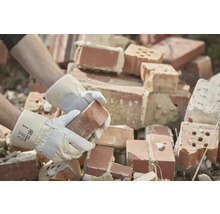 Een persoon met werkhandschoenen houdt een baksteen vast voor een stapel bakstenen.