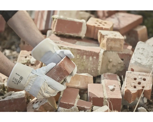 Een persoon met werkhandschoenen houdt een baksteen vast voor een stapel bakstenen.