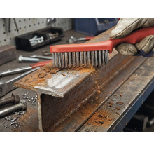 Een hand met werkhandschoen reinigt een roestig stalen profiel met een draadborstel.