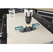 Een boormachine met tegelboorhulp boort in een tegel, een glas water staat ernaast.