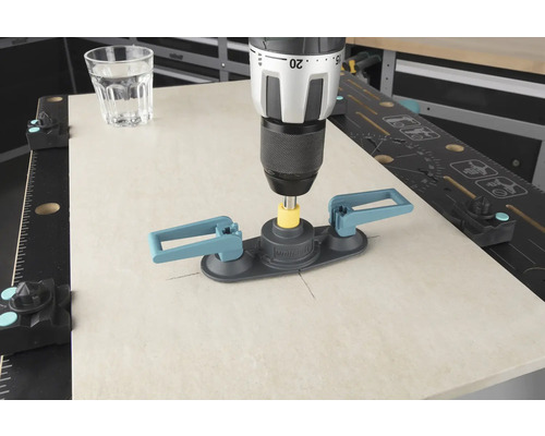 Een boormachine met tegelboorhulp boort in een tegel, een glas water staat ernaast.