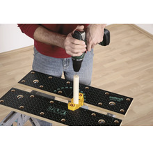 Iemand boort met een boormachine in een stuk hout dat op een werkbank is bevestigd met een geel bevestigingselement.