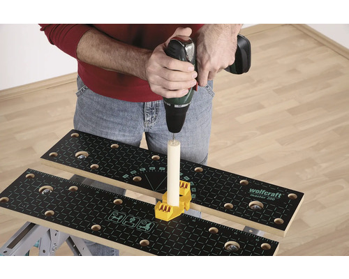 Iemand boort met een boormachine in een stuk hout dat op een werkbank is bevestigd met een geel bevestigingselement.