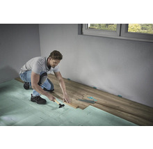 Een man legt laminaatvloeren met een hamer en slagblok op geluiddempende ondervloer in een kamer.