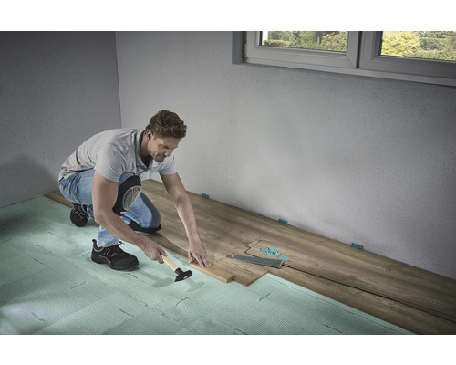 Een man legt laminaatvloeren met een hamer en slagblok op geluiddempende ondervloer in een kamer.