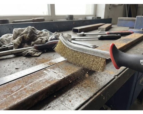 Een draadborstel reinigt een metalen stuk op een werkbank.