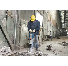 Man met veiligheidshelm en gehoorbescherming bedient een breekhamer in een interieur.