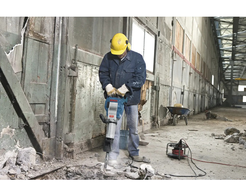 Man met veiligheidshelm en gehoorbescherming bedient een breekhamer in een interieur.