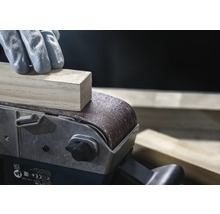 Close-up van een bandschuurmachine die een stuk hout schuurt, bediend door een persoon met handschoenen.