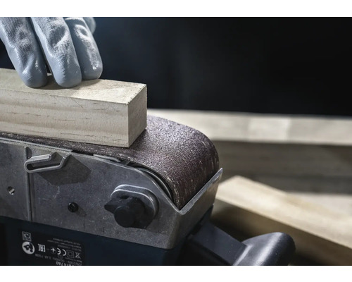 Close-up van een bandschuurmachine die een stuk hout schuurt, bediend door een persoon met handschoenen.