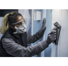 Een persoon draagt beschermende kleding en maakt een muur glad met een gladstrijkspaan.