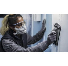 Een persoon draagt beschermende kleding en gebruikt een schuurplank om een muur te schuren.