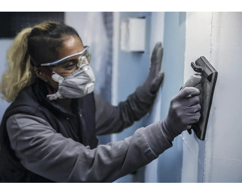 Een persoon draagt beschermende kleding en maakt een muur glad met een gladstrijkspaan.