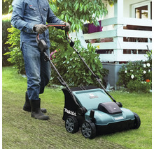 Een persoon met handschoenen en laarzen duwt een Makita verticuteermachine over een gazon in een lichte, goed onderhouden tuin.