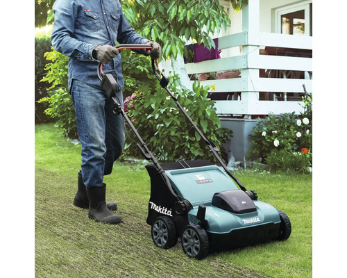 Een persoon met handschoenen en laarzen duwt een Makita verticuteermachine over een gazon in een lichte, goed onderhouden tuin.