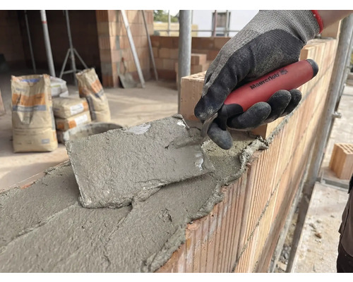 Metselaar werkt met een troffel aan een bakstenen muur, op de achtergrond zakken cement.