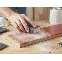 Hand schuurt een gelakte houten plank met een schuurvlies op een schuurblok. Schuurstof is zichtbaar op het hout.