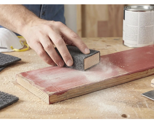 Hand schuurt een gelakte houten plank met een schuurvlies op een schuurblok. Schuurstof is zichtbaar op het hout.