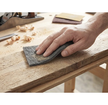 Hand die een houten plank bewerkt met schuurvlies op een lichte werkbank met een houtschaaf en houtkrullen.